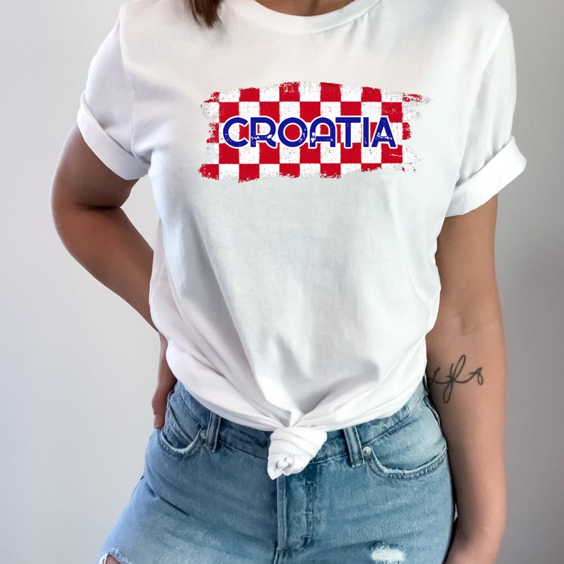 May include: A white short-sleeved t-shirt featuring a rectangular graphic with a red and white checkered pattern. The word CROATIA is printed in blue capital letters across the center of the checkered design, which has a distressed texture.