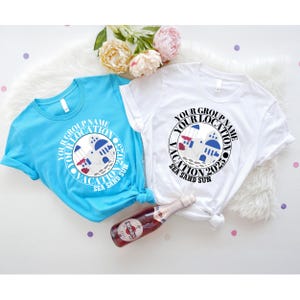 May include: Two short-sleeved t-shirts, one light blue and one white, with a circular graphic. The graphic includes the text "YOUR GROUP NAME YOUR LOCATION VACATION 2025 SEA SAND SUN" around an illustration of a building. A bottle of wine is in the foreground.