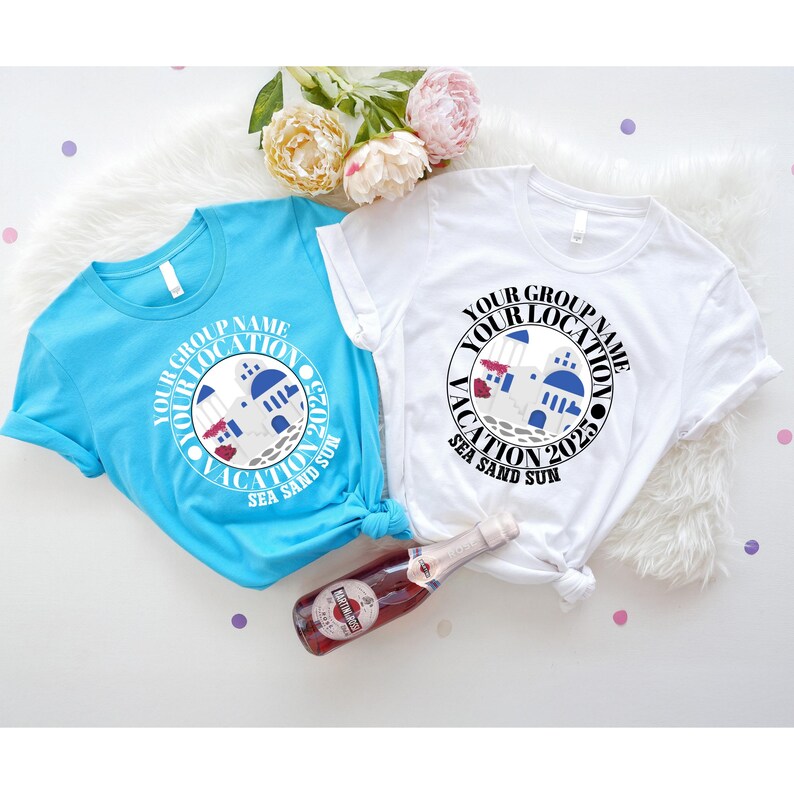 May include: Two short-sleeved t-shirts, one light blue and one white, with a circular graphic. The graphic includes the text "YOUR GROUP NAME YOUR LOCATION VACATION 2025 SEA SAND SUN" around an illustration of a building. A bottle of wine is in the foreground.