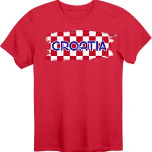 May include: A bright red short-sleeve t-shirt featuring a central graphic of a red and white checkered pattern with a distressed, brush-stroke effect. The word CROATIA is printed in bold, dark blue capital letters across the center of the design.