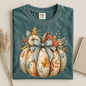 May include: A teal crew neck t-shirt featuring a graphic of a large pumpkin adorned with a blue and white gingham bow. The pumpkin is decorated with various autumn leaves, berries, and flowers. The inner neck label reads Comfort Colors.