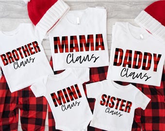 May include: A collection of matching family Christmas pajamas. White t-shirts with "Brother Claus", "Mama Claus", "Daddy Claus", "Mini Claus", and "Sister Claus" in red and black plaid lettering. Red and black plaid pajama pants and red and white Santa hats complete the set.