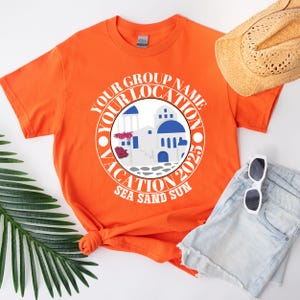 May include: Vibrant orange t-shirt with a circular graphic depicting white buildings with blue domes, and the text "Your Group Name Vacation 2023 Sea Sand Sun". A straw hat, sunglasses, and denim shorts are also visible.