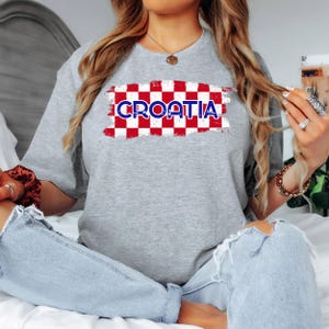 May include: A heather gray short-sleeve t-shirt featuring a central graphic of a red and white checkered pattern with the word CROATIA printed in blue capital letters across the center. The design has a distressed, textured appearance.