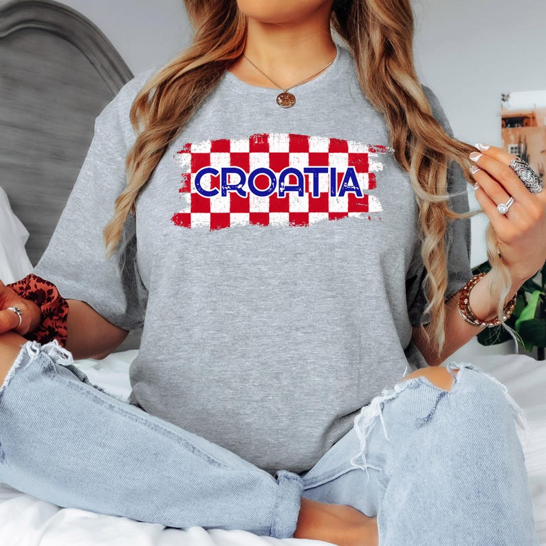 May include: A heather gray short-sleeve t-shirt featuring a central graphic of a red and white checkered pattern with the word CROATIA printed in blue capital letters across the center. The design has a distressed, textured appearance.