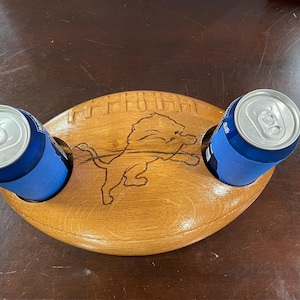 May include: A light brown, wooden, football-shaped drinks holder with two holes for cans. It features a carved Detroit Lions logo and a football design. Two blue cans are placed in the holes.