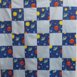 May include: A patchwork quilt featuring a repeating pattern of blue squares with planets, suns, and stars, alternating with solid light blue squares. The planets are orange, yellow, and green. The quilt is likely made of cotton.