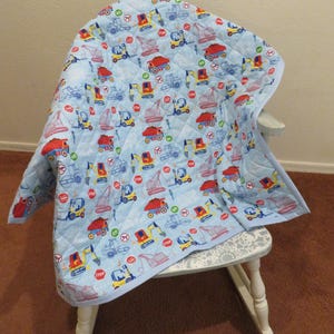 May include: A blue and red baby quilt with a construction theme. The quilt features a variety of construction vehicles, including dump trucks, excavators, and bulldozers. The quilt is quilted with a diamond pattern and has a white border.