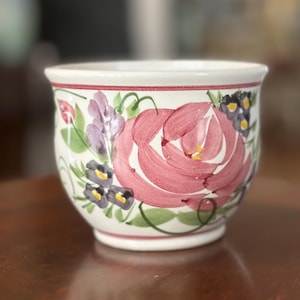 May include: A white ceramic planter with a hand-painted floral design. The design features a large pink rose surrounded by purple and black flowers with green leaves. The rim is trimmed with a red line.
