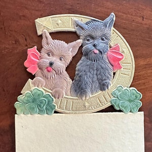 May include: A vintage card with two dogs peeking out from a gold horseshoe. The dogs are wearing pink bows and the horseshoe is decorated with shamrocks. The card has a blank space for writing a message and says "Germany" at the bottom.