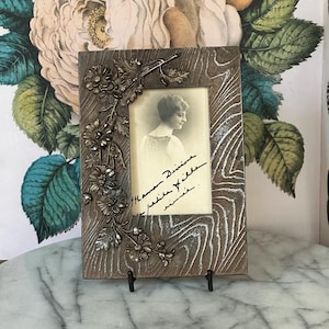 May include: A rectangular wooden picture frame with a black and white photo of a woman. The frame has a decorative floral design on the left side and a wood grain pattern. The photo has handwritten text. The frame is supported by a black metal stand.
