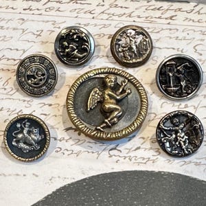 May include: Collection of antique metal buttons with detailed relief designs. The buttons feature various classical motifs, including cherubs, figures, and heraldic symbols. The buttons are in shades of silver and gold, with intricate detailing.