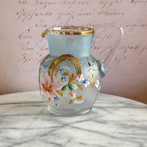 May include: A small, frosted blue glass pitcher with a gold rim and painted floral design. The pitcher has a clear glass handle and is decorated with pink, white, and yellow flowers, as well as a gold horseshoe.