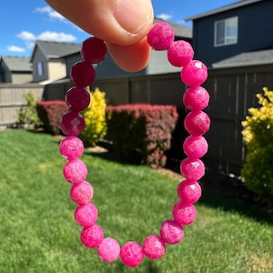 Könnte beinhalten: Ein elastisches Armband bestehend aus zahlreichen runden, facettierten Perlen in einem leuchtenden, durchscheinenden Pink. Die Perlen sind in Größe und Form einheitlich und zu einer kreisförmigen Schlaufe aufgereiht, die das Sonnenlicht einfängt.