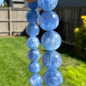 Könnte beinhalten: Ein Armband aus runden, hellblauen Kyanit-Perlen. Die Perlen haben eine durchscheinende Qualität und sind aufgefädelt. Das Armband wird vor einem Hintergrund aus grünem Gras und blauem Himmel gehalten.
