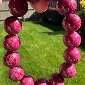 May include: A beaded bracelet featuring large, spherical, polished magenta-colored stones with natural surface textures and dark inclusions. The beads are strung together in a circular loop, showcasing a vibrant, deep pink hue throughout the piece.