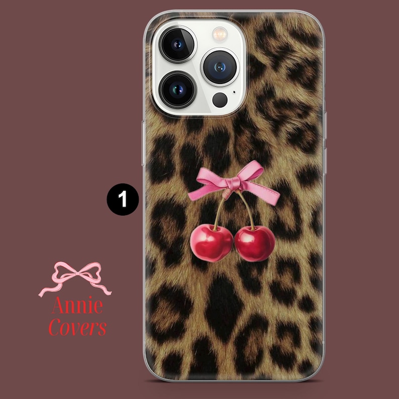 Puede incluir: Funda transparente para tel&eacute;fono con dise&ntilde;o de estampado de leopardo, un lazo rosa y dos cerezas rojas. La funda tiene un borde transparente y un recorte para la c&aacute;mara. El texto "Annie Covers" es visible en la esquina inferior izquierda.
