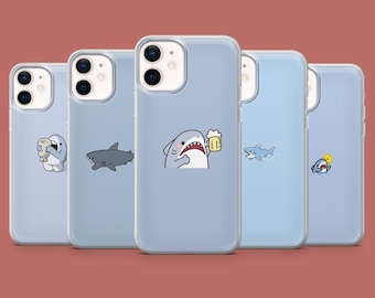 May include: Five light blue phone cases with various shark-themed illustrations. One case features a shark holding a beer, another a shark with a seal, and others with shark designs. The cases are arranged against a solid red-brown background.