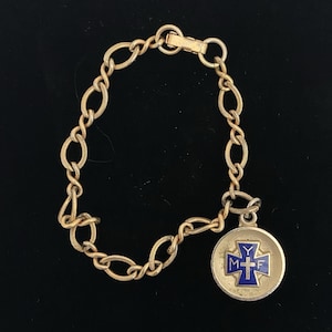 May include: A gold-toned metal chain bracelet featuring a round pendant with a blue enamel cross. The cross displays the letters Y, M, and F, along with a small central cross symbol. The bracelet includes a secure clasp mechanism for closure.