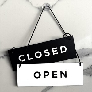 Op de afbeelding: Een zwart-wit bord met de woorden "GESLOTEN" en "OPEN" hangend aan een touwtje. Het bord is gemaakt van twee rechthoekige stukken karton, een zwart en een wit.