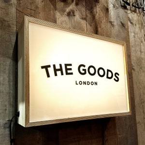 Pode incluir: Uma placa de caixa de luz branca com uma moldura de madeira preta. A placa diz "THE GOODS LONDON" em texto preto.