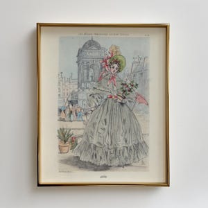 Puede incluir: Una ilustración vintage de una mujer con un vestido gris y un sombrero verde, sosteniendo un ramo de flores. La ilustración está enmarcada en oro y presenta el texto "Les Modes Parisiennes du Temps Présent" y "J.A.S.B."