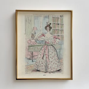 Peut inclure: Une illustration vintage encadrée d'une femme en robe fleurie avec une ceinture rose. La femme se tient dans une pièce avec un piano et une bibliothèque. L'illustration est intitulée "Les Modes Parisiennes de 1850".