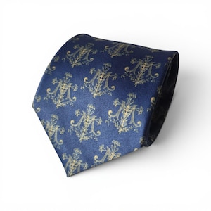 May include: A dark blue necktie featuring a repeating pattern of gold-colored letter M monograms surrounded by intricate floral and botanical illustrations. The tie has a smooth, satin-like finish and a pointed tip, displaying a consistent design.
