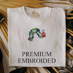 Op de afbeelding: Wit T-shirt met een kleurrijk geborduurd rupsontwerp en de tekst "EAT THE RICH". Het shirt heeft ook de woorden "PREMIUM EMBROIDERED" eronder gedrukt. Het shirt wordt gepresenteerd met accessoires.