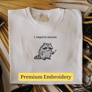 May include: White t-shirt with the text "I require snacks" above a cartoon raccoon holding a knife. The shirt has "Premium Embroidery" in a yellow rectangle at the bottom. The shirt is on a neutral background with accessories.