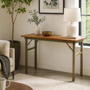 Può includere: Una consolle in legno con struttura in metallo verde anticato. Il piano del tavolo è marrone caldo e le gambe hanno un aspetto vintage. Una lampada bianca, una piccola ciotola di ottone e un vaso bianco con piante verdi sono sul tavolo. Una stampa floreale incorniciata è appesa al muro.