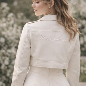 Könnte beinhalten: Elfenbeinfarbene, kurze Lederjacke mit dem gestickten Namen "Mrs. Anderson" auf dem Rücken. Die Jacke hat einen klassischen Biker-Stil mit Reverskragen, Reißverschlussdetails und einer taillierten Silhouette, ideal für Hochzeiten.