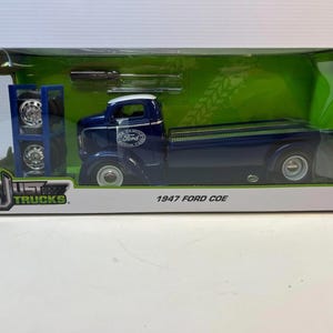 May include: A die-cast model of a 1947 Ford COE truck in a display box. The truck is dark blue with white accents and the Ford logo. The box includes accessories and additional wheels.