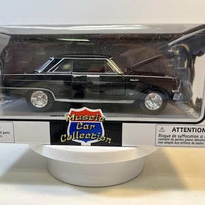 May include: A 1:25 scale die-cast black muscle car model inside a clear plastic display box. The packaging features the text Muscle Car Collection, a warning label, and an age recommendation of 5+ years. The car has silver wheels and trim.