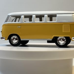 May include: A die-cast scale model of a vintage van featuring a two-tone color scheme with a mustard yellow lower body and a white roof. The vehicle includes silver-colored hubcaps, clear windows, and a rectangular vent on the rear side panel.