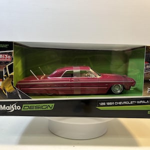 May include: A 1:26 scale die-cast metal 1964 Chevrolet Impala SS model car in a magenta finish with white pinstriping and chrome wire wheels. The vehicle is enclosed in a Maisto Design Lowriders branded display box with clear plastic window.