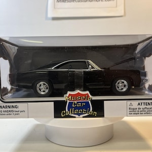 May include: A black 1:25 scale die-cast muscle car model inside a rectangular display box. The packaging features the text Muscle Car Collection, NewRay branding, and safety warnings. The car has chrome-colored wheels and a visible engine bay.