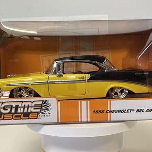 May include: A yellow and black 1956 Chevrolet Bel Air die-cast model car by Jada Toys, displayed inside its original Big Time Muscle packaging. The vehicle features chrome wheels, a two-tone paint scheme, and clear plastic window casing.