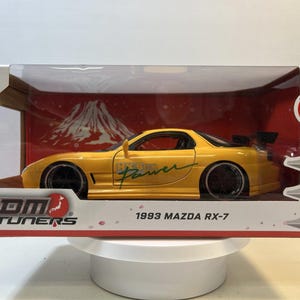 May include: A die-cast 1993 Mazda RX-7 model car in a clear display case. The car is yellow with black wheels and the words "Hyper Power" on the side. The display case has the JDM Tuners logo and a graphic of a mountain.