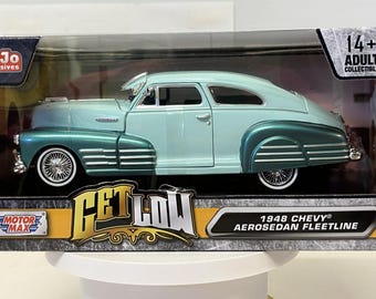 May include: A 1948 Chevy Aerosedan Fleetline die-cast model car in a two-tone light blue and teal finish. The vehicle features wire-spoke wheels with white-wall tires, chrome trim, and is packaged in a box labeled Get Low and 14+ Adult Collectible.