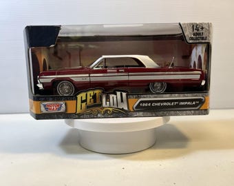 May include: A 1964 Chevrolet Impala die-cast model car in deep red with a white side stripe, featuring wire-spoke wheels. The vehicle is housed in a rectangular display box labeled Get Low and Motor Max, with a clear plastic window for viewing.