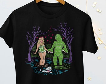 Könnte beinhalten: Schwarzes T-Shirt mit einem Grafikdruck einer Frau und einer Kreatur aus der Schwarzen Lagune, die sich in einem Sumpf die Hände halten. Die Frau trägt einen grün-weißen Badeanzug und die Kreatur ist grün. Über ihnen schweben Herzen.