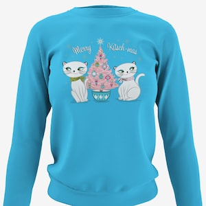 Puede incluir: Una sudadera de manga larga azul turquesa con un diseño navideño que presenta dos gatos blancos a ambos lados de un árbol de Navidad rosa con adornos. El texto "Merry Kitsch-mas" está impreso sobre el árbol.