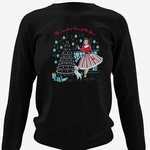 Puede incluir: Sudadera negra de manga larga con una escena navideña retro que presenta a una mujer con un vestido rojo y un perro blanco decorando un árbol de Navidad. El texto "The Loveliest Time of the Year" está impreso sobre la escena.