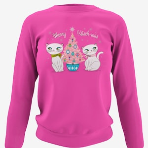 Puede incluir: Sudadera rosa de manga larga con un árbol de Navidad y dos gatos con bufandas blancas. El texto "Merry Kitsch-mas" está impreso en la sudadera.