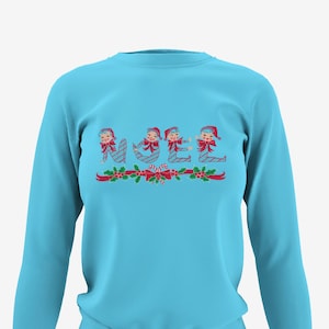 Puede incluir: Una sudadera de manga larga azul claro con un diseño navideño que presenta la palabra "NOEL" deletreada con duendes que llevan atuendos a rayas de bastón de caramelo y lazos rojos. Los duendes están rodeados de acebo y bastones de caramelo.