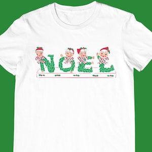 Puede incluir: Camiseta blanca con un diseño navideño que representa la palabra "NOEL" escrita con cinco duendes que llevan coronas de acebo.