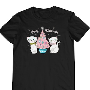 Puede incluir: Camiseta negra con un árbol de Navidad rosa y dos gatos blancos. El texto "Merry Kitsch-mas" está por encima del árbol.