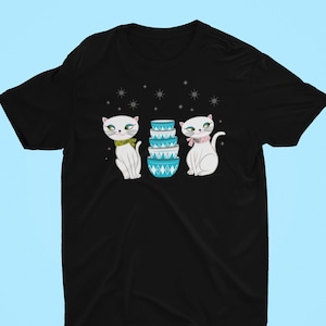 Puede incluir: Camiseta negra con un gráfico de dos gatos blancos con ojos azules y narices rosas, de pie a ambos lados de una pila de tazones de mezclar azules y blancos. El fondo es negro con copos de nieve blancos.