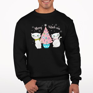 Puede incluir: Sudadera negra con un árbol de Navidad blanco y dos gatos blancos con lazos rosas. El texto "Merry Kitsch-mas" está impreso en la sudadera.